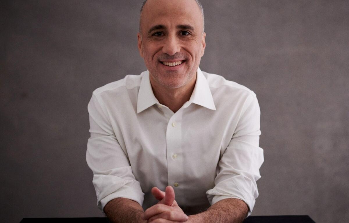 Smiling middle-aged man in a white dress shirt sits at a table with hands clasped in front of him against a gray backdrop.