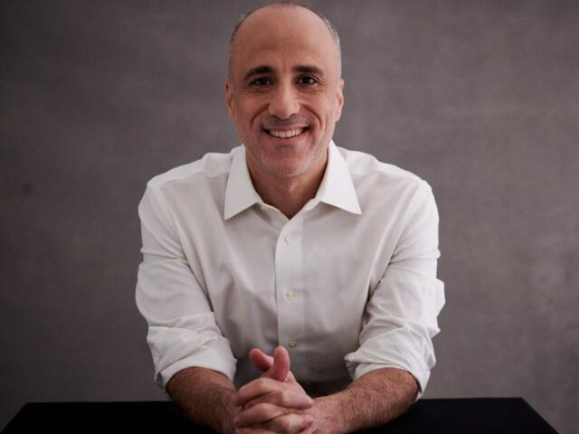 Smiling middle-aged man in a white dress shirt sits at a table with hands clasped in front of him against a gray backdrop.