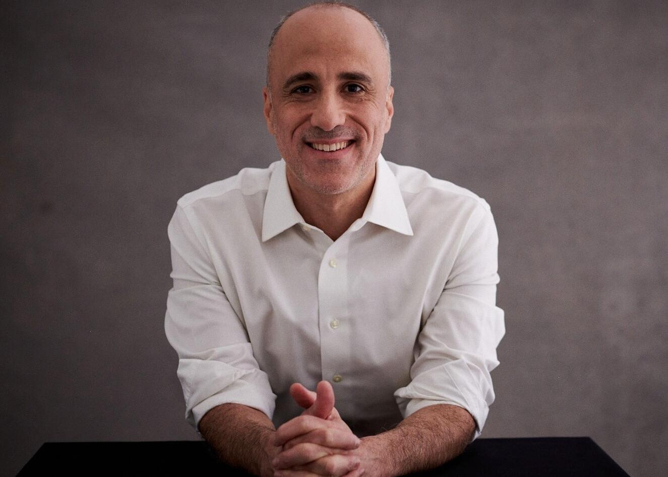 Smiling middle-aged man in a white dress shirt sits at a table with hands clasped in front of him against a gray backdrop.