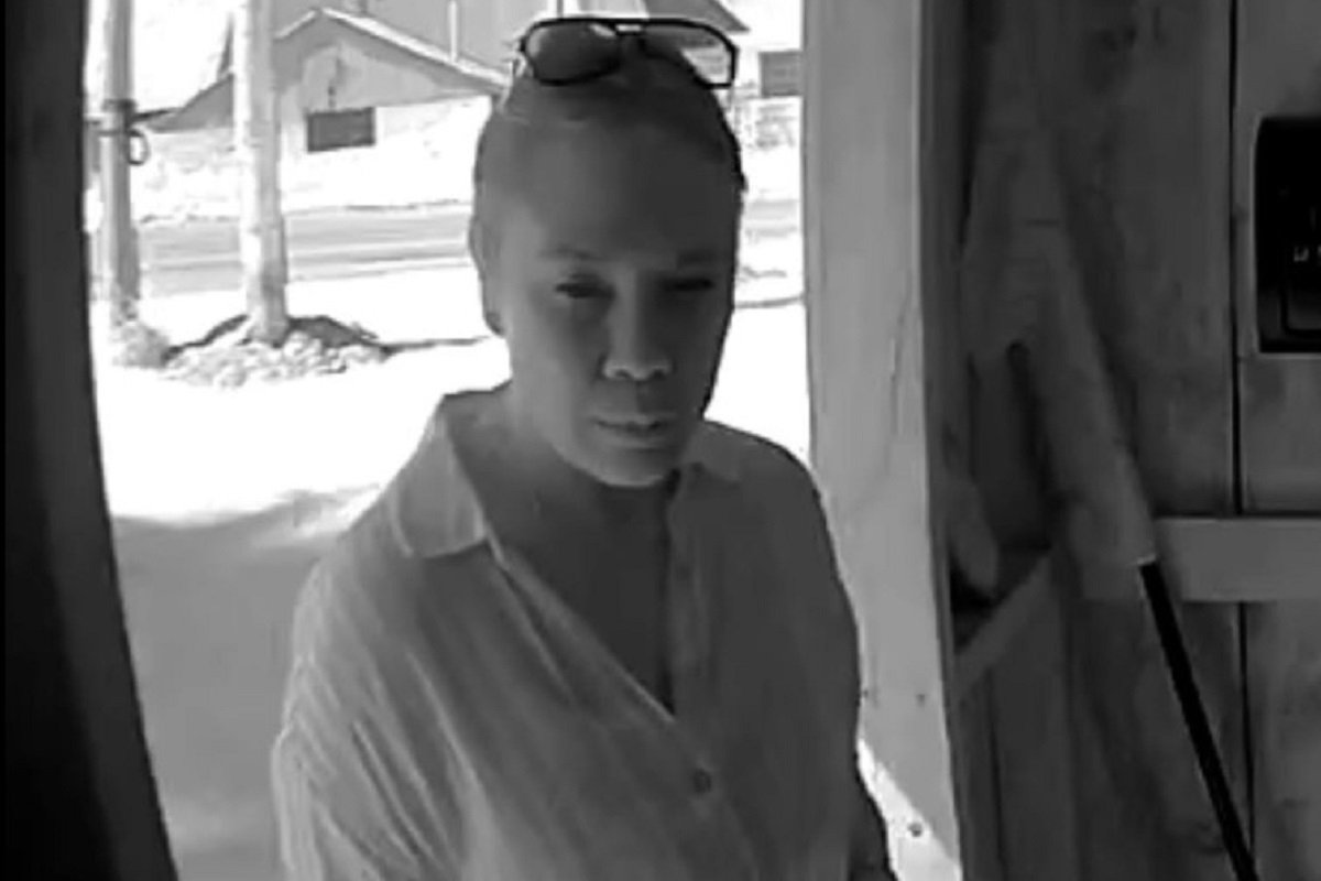 Woman in a light shirt stands at a doorway, sunglasses on her head, looking toward the camera.