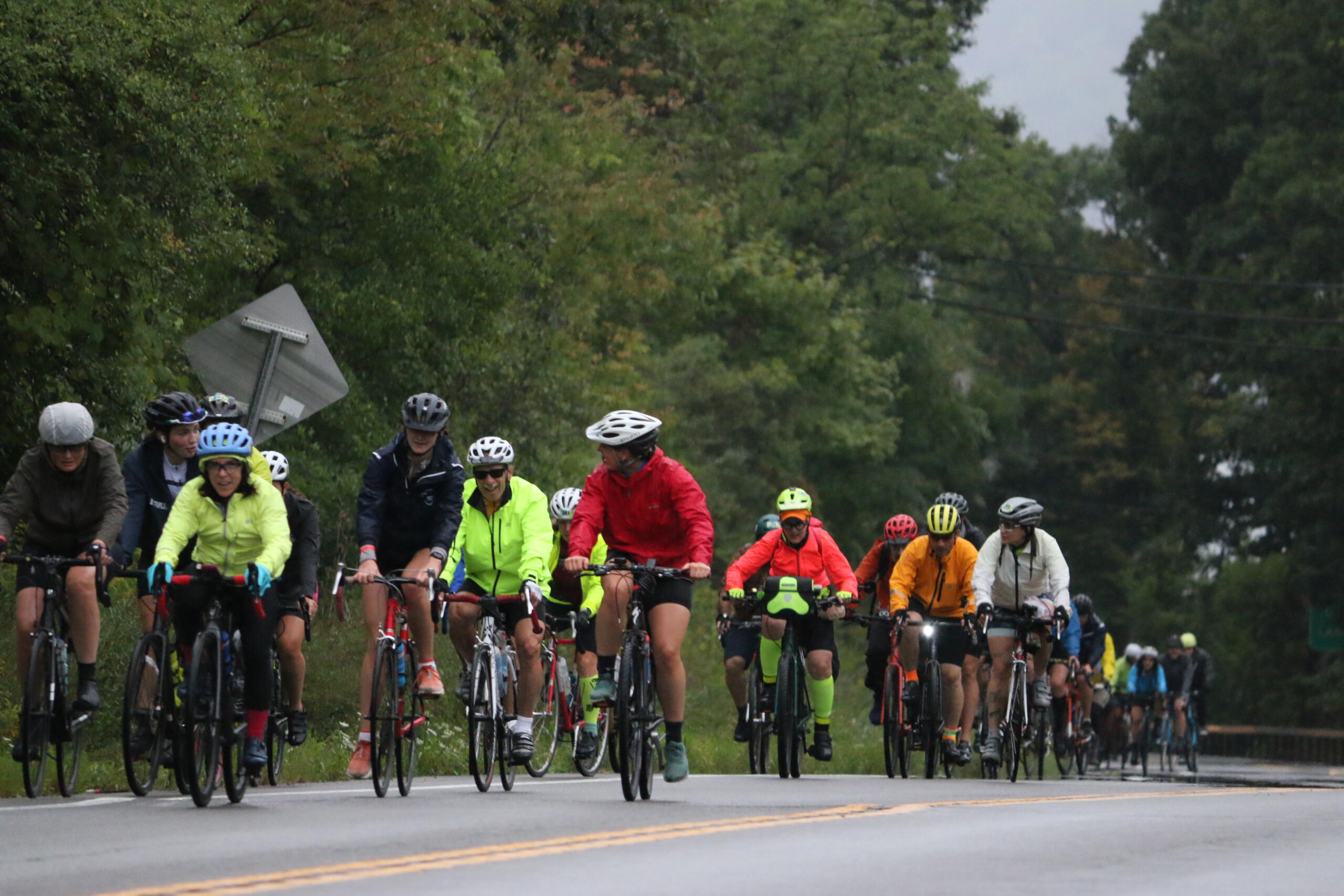Group of cyclists wearing colorful jackets and helmets riding on a two-lane road with trees on both sides.