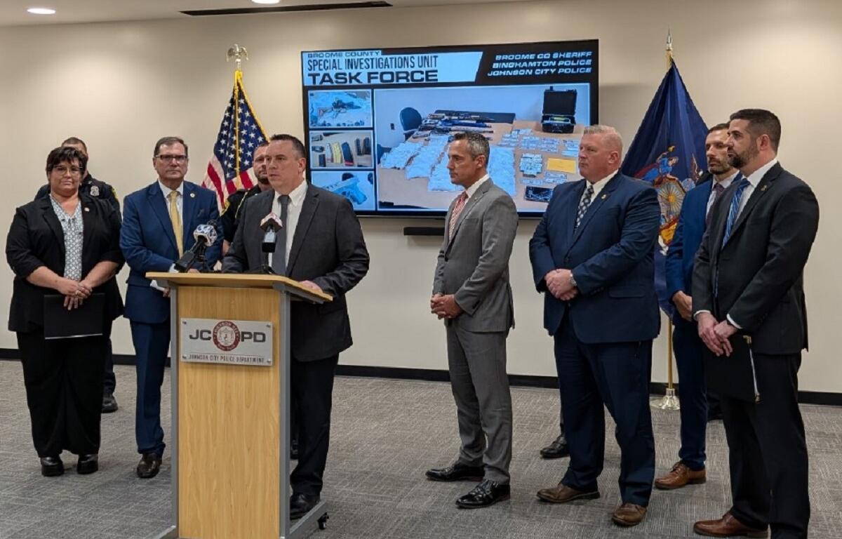 A man in a dark suit speaks at a wooden podium during a police press conference, with six other officials in suits standing behind him; flags flank the group and a large screen labeled 'Task Force' shows seized items.