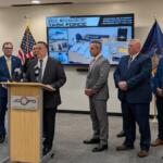 A man in a dark suit speaks at a wooden podium during a police press conference, with six other officials in suits standing behind him; flags flank the group and a large screen labeled 'Task Force' shows seized items.