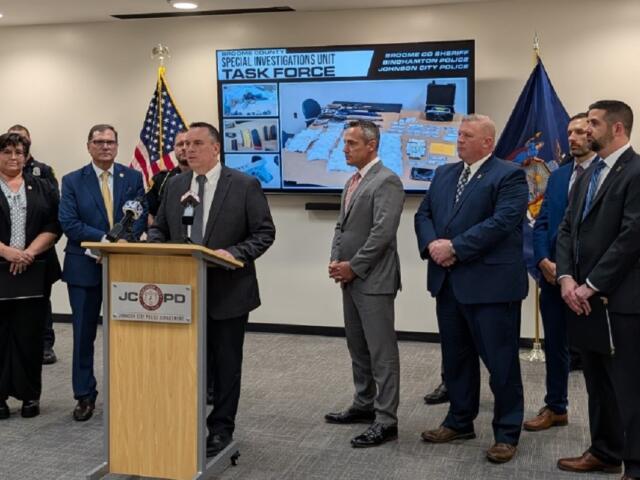 A man in a dark suit speaks at a wooden podium during a police press conference, with six other officials in suits standing behind him; flags flank the group and a large screen labeled 'Task Force' shows seized items.