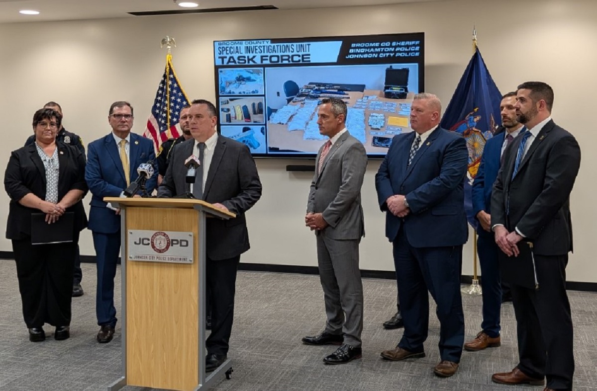 A man in a dark suit speaks at a wooden podium during a police press conference, with six other officials in suits standing behind him; flags flank the group and a large screen labeled 'Task Force' shows seized items.