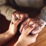 Young adult and elderly person hold hands on a wooden table, conveying support and care.