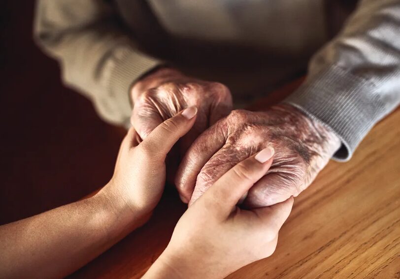 Young adult and elderly person hold hands on a wooden table, conveying support and care.