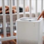Hand pressing the top control of a white air purifier in a baby's nursery with a crib in the background.