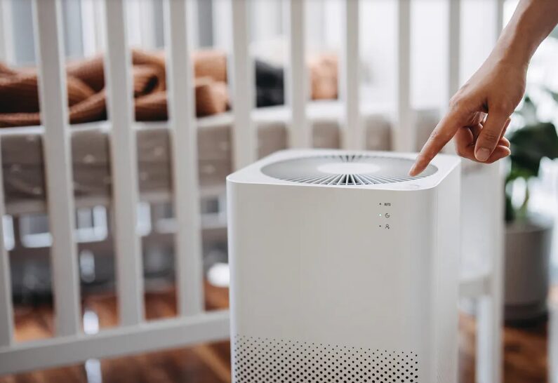 Hand pressing the top control of a white air purifier in a baby's nursery with a crib in the background.