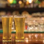 Two tall pint glasses filled with golden beer sit on a wooden bar, blurred bar lights in the background.