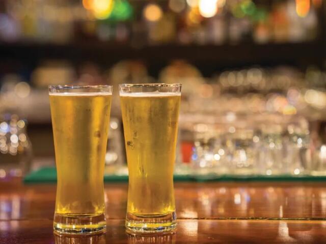 Two tall pint glasses filled with golden beer sit on a wooden bar, blurred bar lights in the background.