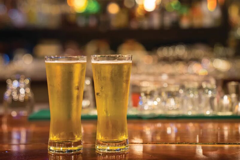 Two tall pint glasses filled with golden beer sit on a wooden bar, blurred bar lights in the background.