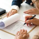 Two people sign a contract on a clipboard at a white desk, with a pen in one hand and the document held steady by the other.