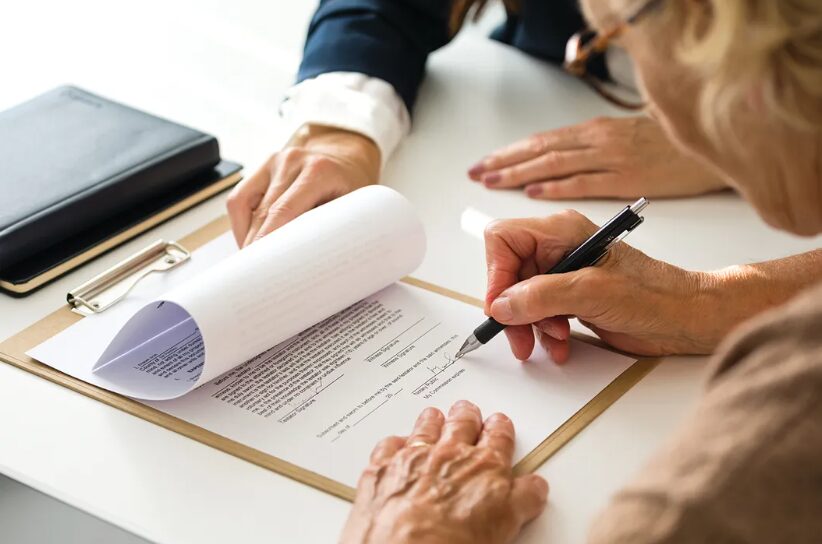 Two people sign a contract on a clipboard at a white desk, with a pen in one hand and the document held steady by the other.