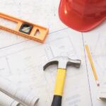 Construction tools and blueprints on a drafting table: hammer, level, pencils, rolls, and a red hard hat nearby