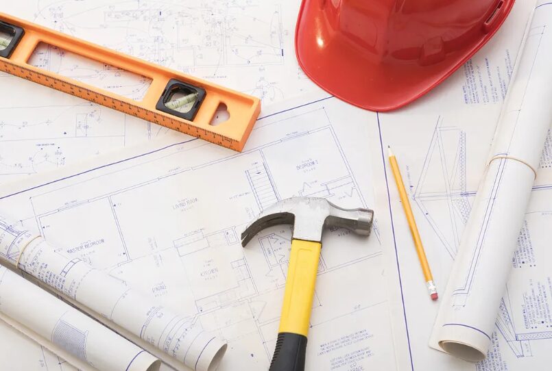 Construction tools and blueprints on a drafting table: hammer, level, pencils, rolls, and a red hard hat nearby