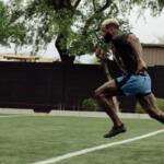 Male sprinter mid-stride on a turf field, wearing a black shirt and blue shorts, with orange cones in the background.