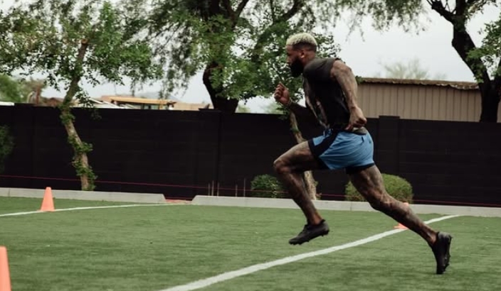 Male sprinter mid-stride on a turf field, wearing a black shirt and blue shorts, with orange cones in the background.