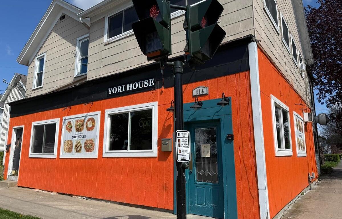 Front view of Yori House restaurant, a bright orange building with a teal door and menu posters in the windows, on a sunny day.