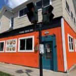 Front view of Yori House restaurant, a bright orange building with a teal door and menu posters in the windows, on a sunny day.