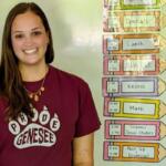 Smiling female student in a maroon shirt standing next to a colorful classroom schedule display showing periods like Lunch and Math.