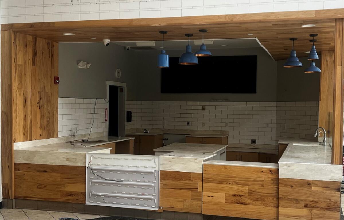 Empty cafe counter area with wood panels, marble-like countertops, white subway tiles, and blue pendant lights.