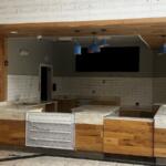 Empty cafe counter area with wood panels, marble-like countertops, white subway tiles, and blue pendant lights.