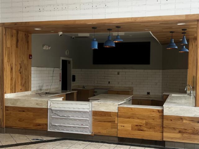 Empty cafe counter area with wood panels, marble-like countertops, white subway tiles, and blue pendant lights.