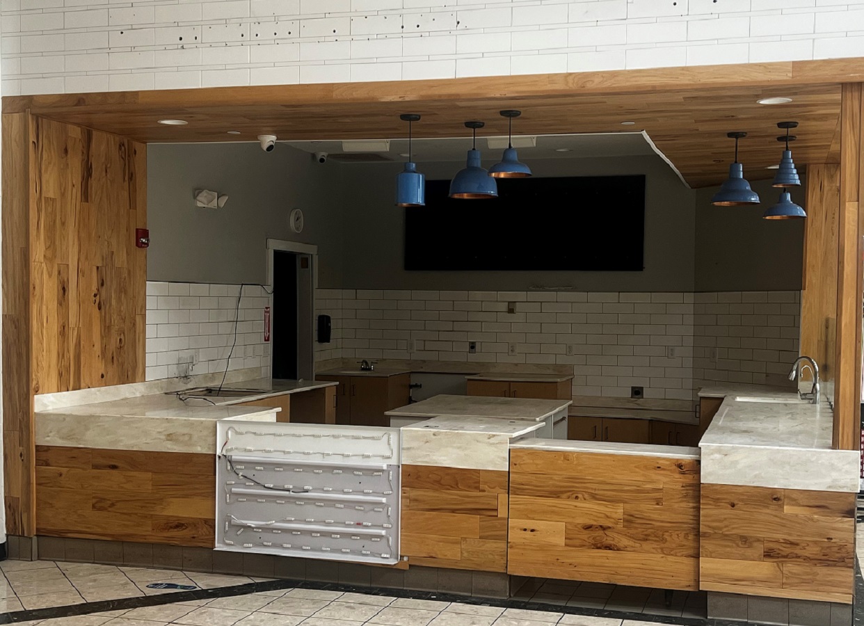 Empty cafe counter area with wood panels, marble-like countertops, white subway tiles, and blue pendant lights.
