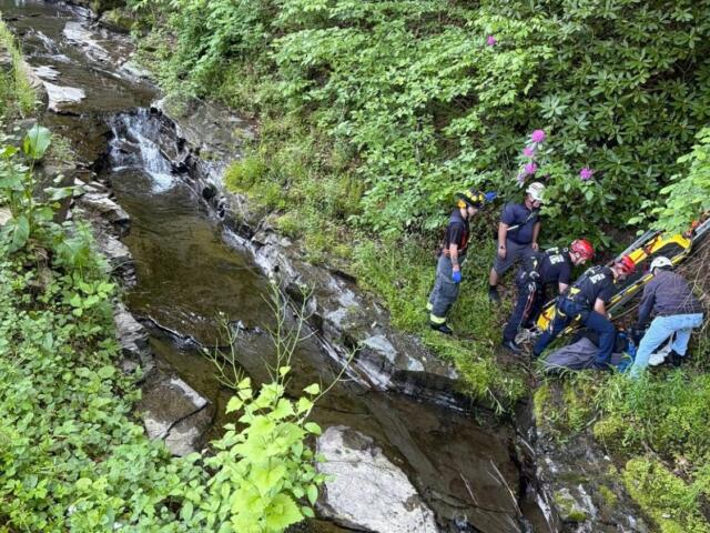 Emergency responders carry a patient on a stretcher up a rocky, overgrown riverbank.