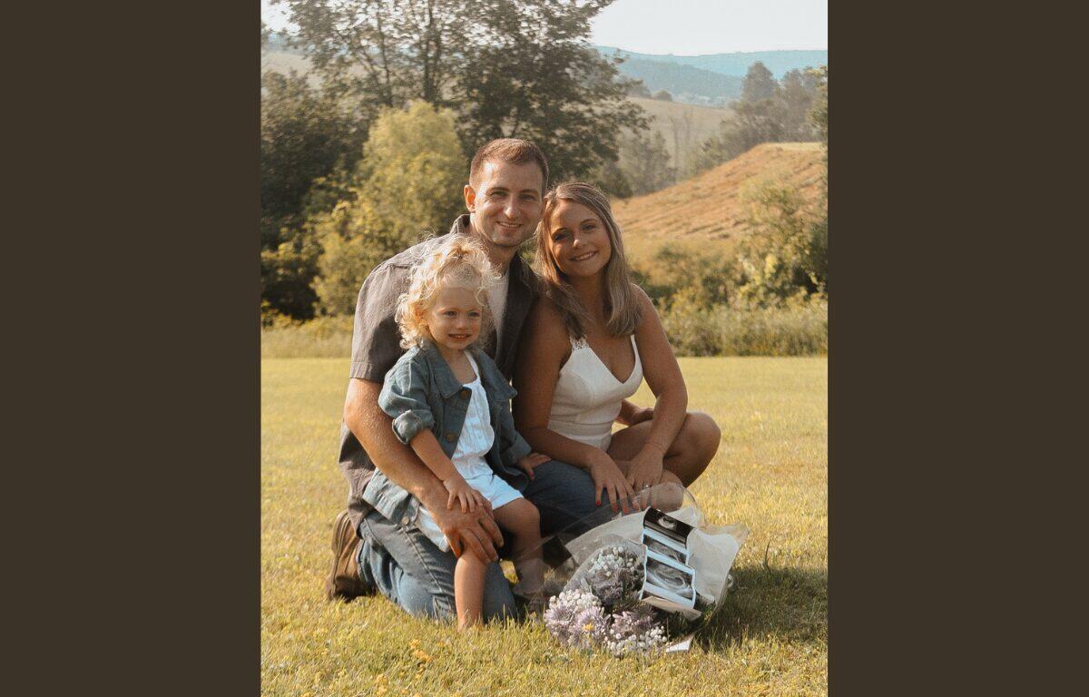 Family of three sitting on a sunny grassy field, smiling at the camera with flowers nearby.