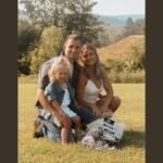 Family of three sitting on a sunny grassy field, smiling at the camera with flowers nearby.