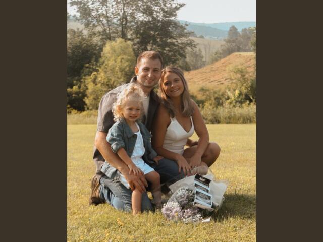 Family of three sitting on a sunny grassy field, smiling at the camera with flowers nearby.