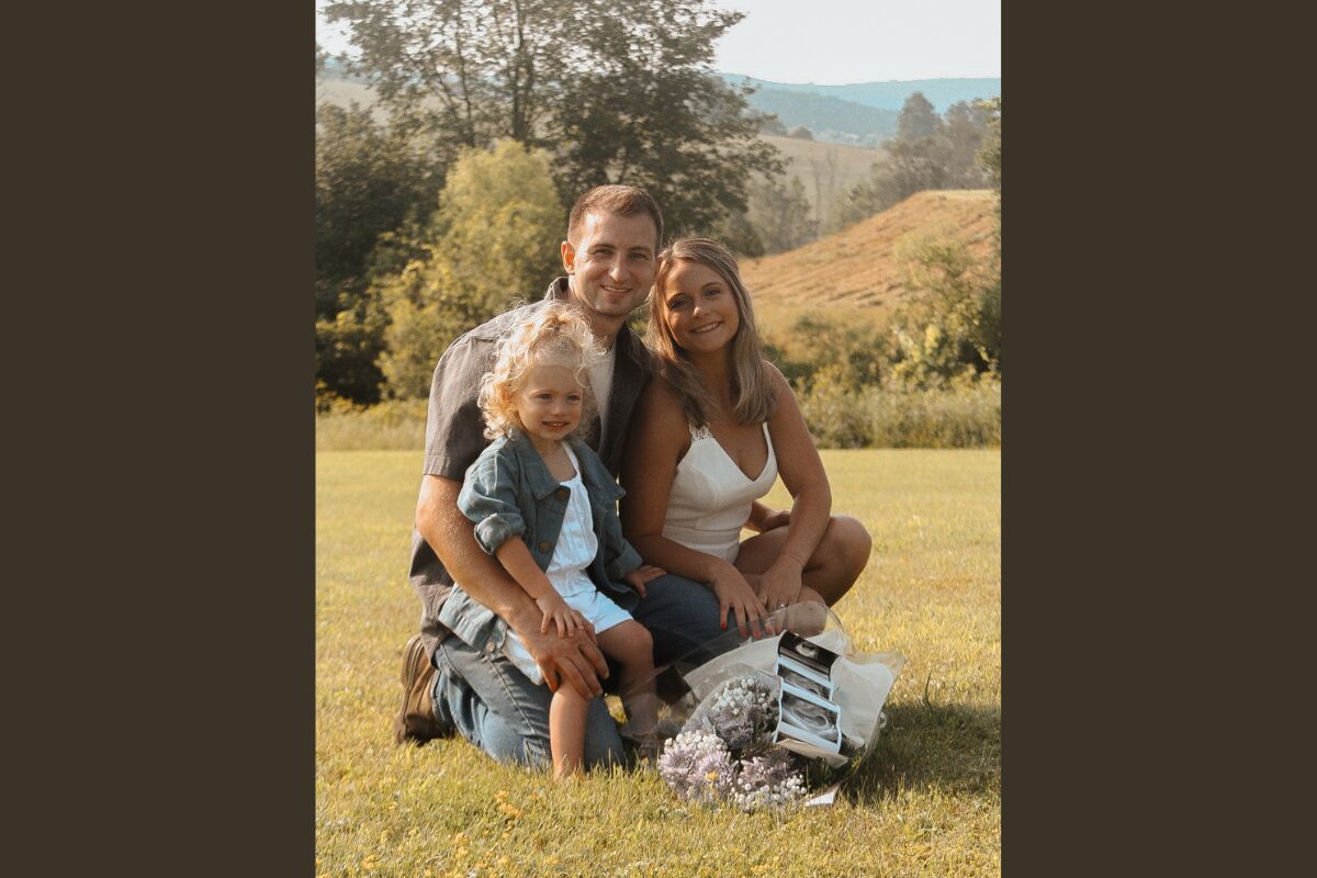 Family of three sitting on a sunny grassy field, smiling at the camera with flowers nearby.