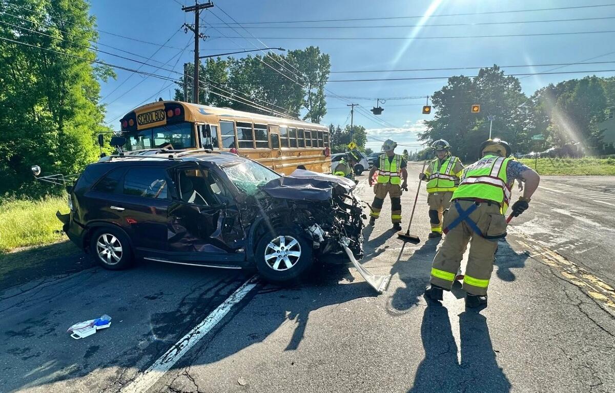 Front-end crash involving a black SUV and a yellow school bus; firefighters in reflective gear respond at a sunny intersection on a suburban road.