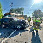 Front-end crash involving a black SUV and a yellow school bus; firefighters in reflective gear respond at a sunny intersection on a suburban road.