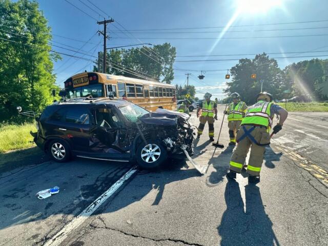 Front-end crash involving a black SUV and a yellow school bus; firefighters in reflective gear respond at a sunny intersection on a suburban road.