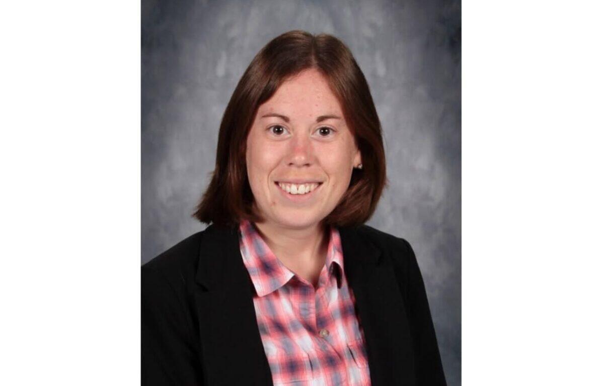 Smiling woman in a black blazer and pink plaid shirt, head-and-shoulders portrait against a gray studio backdrop.