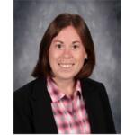 Smiling woman in a black blazer and pink plaid shirt, head-and-shoulders portrait against a gray studio backdrop.