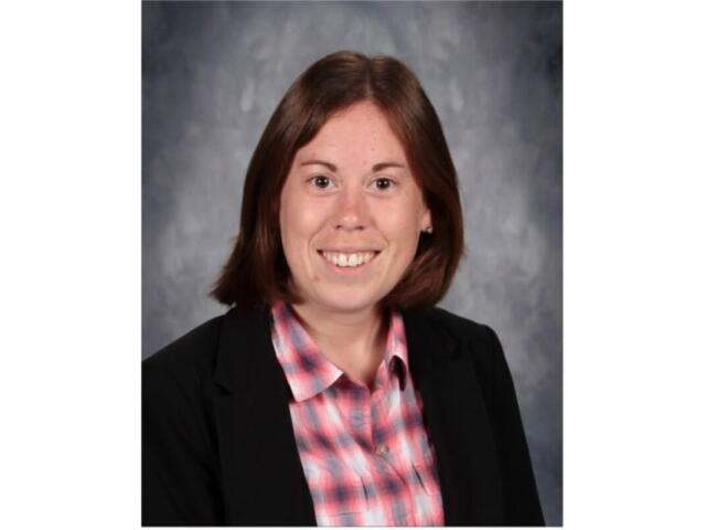 Smiling woman in a black blazer and pink plaid shirt, head-and-shoulders portrait against a gray studio backdrop.