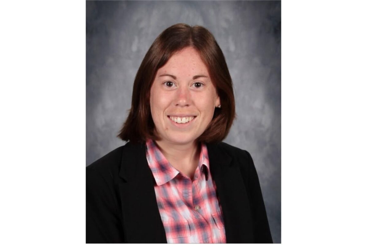 Smiling woman in a black blazer and pink plaid shirt, head-and-shoulders portrait against a gray studio backdrop.