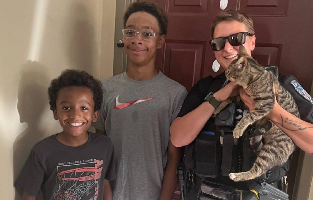 Police officer in uniform holding a tabby cat, posing with two smiling boys at a doorway.