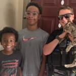 Police officer in uniform holding a tabby cat, posing with two smiling boys at a doorway.