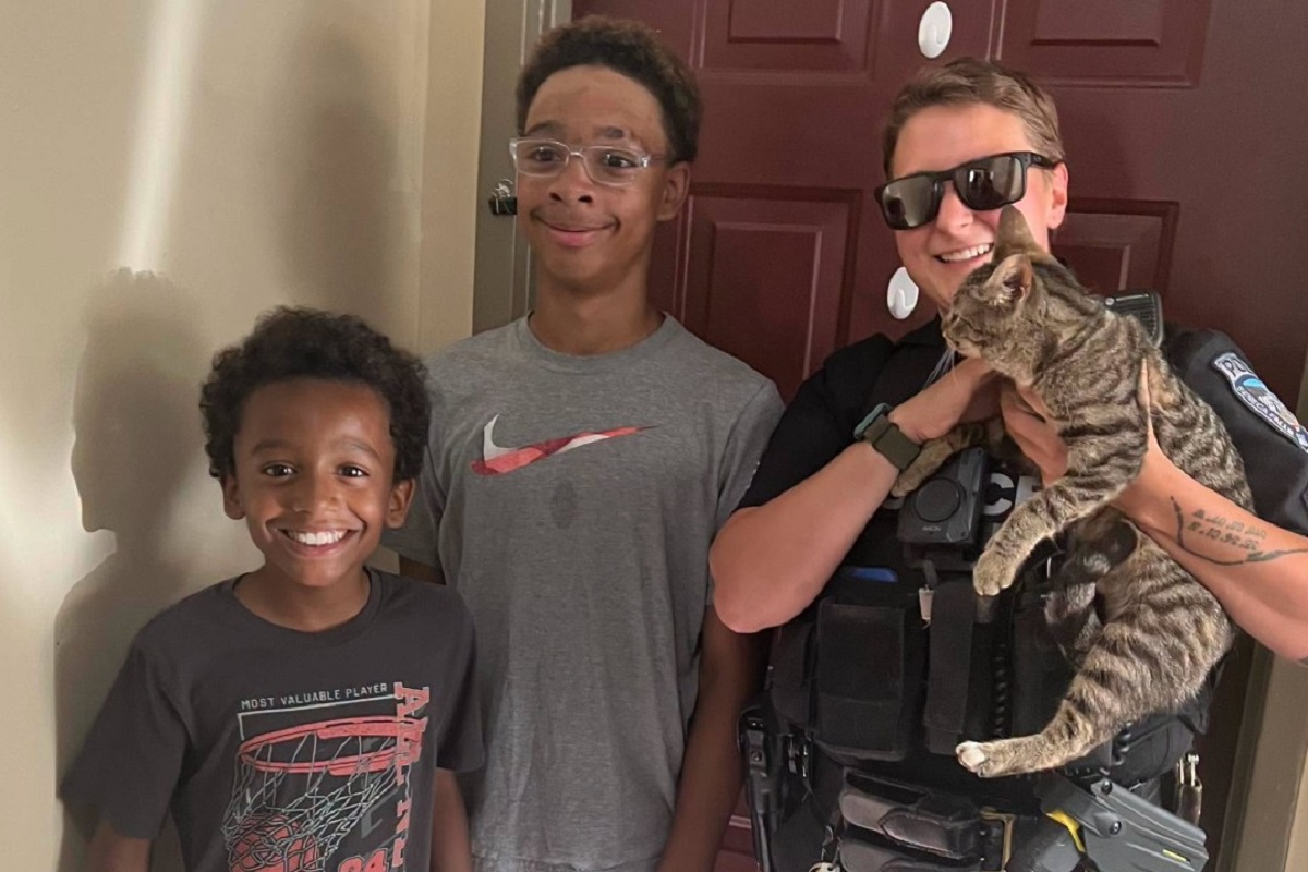 Police officer in uniform holding a tabby cat, posing with two smiling boys at a doorway.