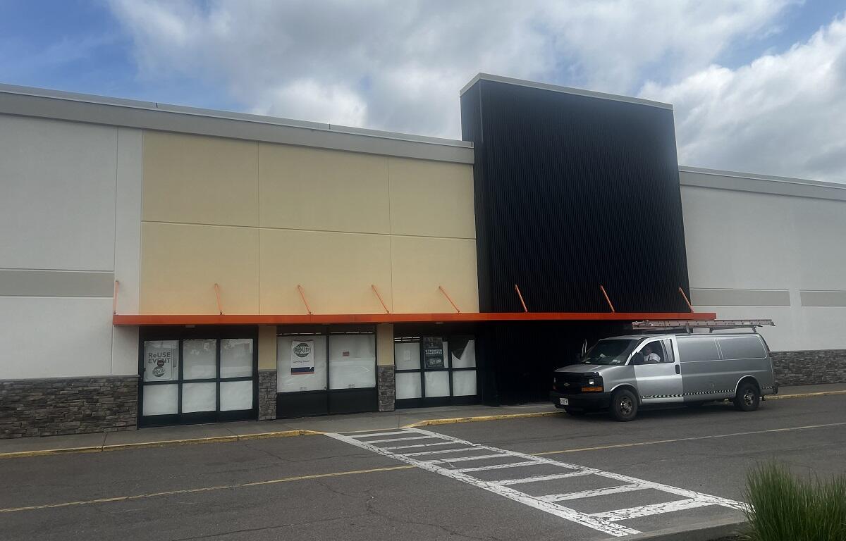 Exterior of a retail storefront with an orange awning, beige and black facade, and a silver van parked in front.