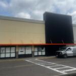 Exterior of a retail storefront with an orange awning, beige and black facade, and a silver van parked in front.