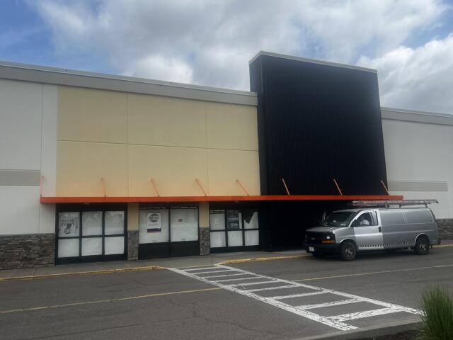 Exterior of a retail storefront with an orange awning, beige and black facade, and a silver van parked in front.