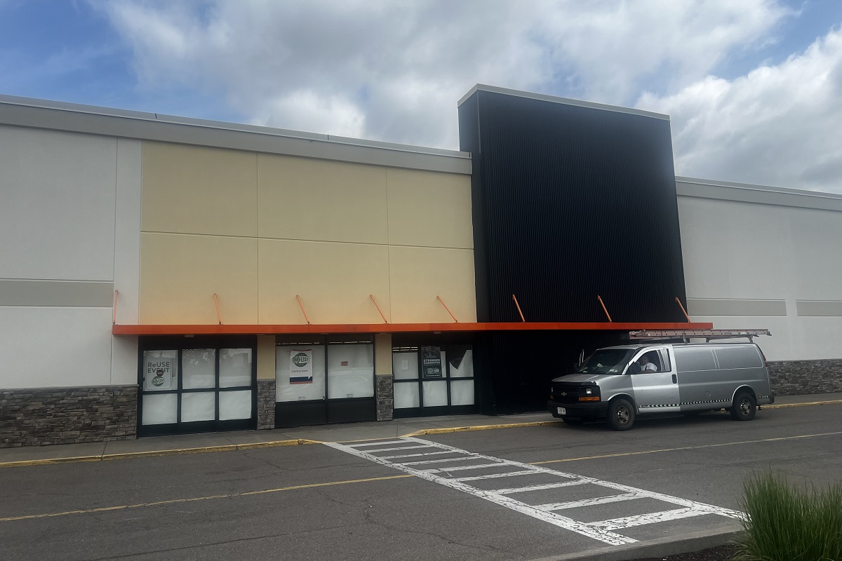 Exterior of a retail storefront with an orange awning, beige and black facade, and a silver van parked in front.