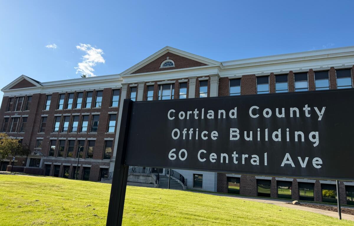 Brick office building with white trim and arched window, under a blue sky; foreground sign reads Cortland County Office Building, 60 Central Ave (partial).