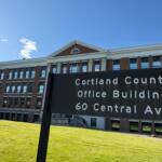 Brick office building with white trim and arched window, under a blue sky; foreground sign reads Cortland County Office Building, 60 Central Ave (partial).